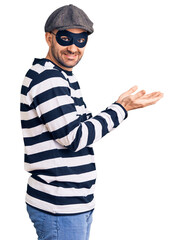 Poster - Young handsome man wearing burglar mask pointing aside with hands open palms showing copy space, presenting advertisement smiling excited happy