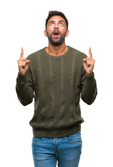 Canvas Print - Adult hispanic man wearing winter sweater over isolated background amazed and surprised looking up and pointing with fingers and raised arms.