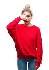 Wall Mural - Young beautiful blonde woman wearing red sweater and glasses over isolated background peeking in shock covering face and eyes with hand, looking through fingers with embarrassed expression.