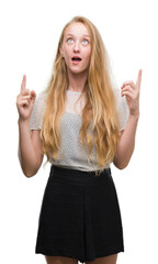 Poster - Blonde teenager woman wearing moles shirt amazed and surprised looking up and pointing with fingers and raised arms.