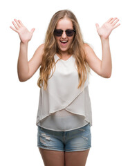 Wall Mural - Young blonde woman wearing sunglasses celebrating mad and crazy for success with arms raised and closed eyes screaming excited. Winner concept