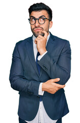 Wall Mural - Hispanic man with beard wearing business suit and tie serious face thinking about question with hand on chin, thoughtful about confusing idea