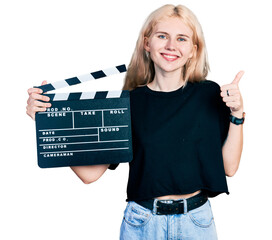 Poster - Young caucasian woman holding video film clapboard smiling happy and positive, thumb up doing excellent and approval sign