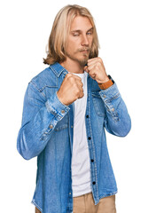 Wall Mural - Caucasian man with blond long hair wearing casual denim jacket ready to fight with fist defense gesture, angry and upset face, afraid of problem