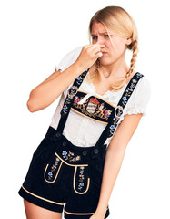 Wall Mural - Young beautiful blonde woman wearing oktoberfest dress smelling something stinky and disgusting, intolerable smell, holding breath with fingers on nose. bad smell