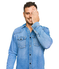 Sticker - Young hispanic man wearing casual denim jacket covering one eye with hand, confident smile on face and surprise emotion.