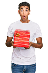 Wall Mural - Young handsome african american man holding gift scared and amazed with open mouth for surprise, disbelief face