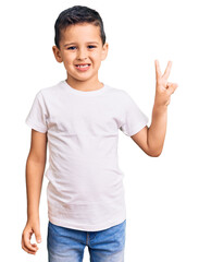 Wall Mural - Little cute boy kid wearing casual white tshirt smiling with happy face winking at the camera doing victory sign. number two.