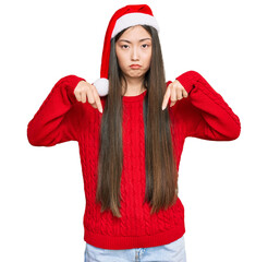 Canvas Print - Young chinese woman wearing christmas hat pointing down looking sad and upset, indicating direction with fingers, unhappy and depressed.