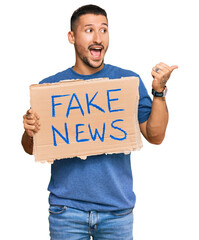 Sticker - Handsome man with tattoos holding fake news banner pointing thumb up to the side smiling happy with open mouth