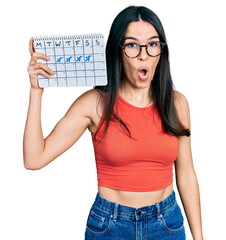Young hispanic woman holding travel calendar scared and amazed with open mouth for surprise, disbelief face
