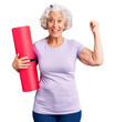© Krakenimages.com - Senior grey-haired woman holding yoga mat screaming proud, celebrating victory and success very excited with raised arms