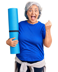 Poster - Senior woman with gray hair holding yoga mat screaming proud, celebrating victory and success very excited with raised arms