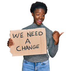 Canvas Print - Young african american girl holding we need a change banner pointing thumb up to the side smiling happy with open mouth