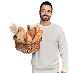 Poster - Young hispanic man with beard holding wicker basket with bread looking positive and happy standing and smiling with a confident smile showing teeth