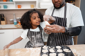 Happy African American kid girl and father cooking break or bakery with rolling bread in kitchen at home	