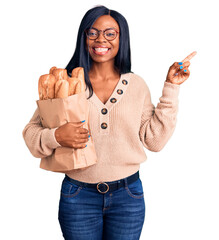 Wall Mural - Young african american woman holding paper bag with bread smiling happy pointing with hand and finger to the side