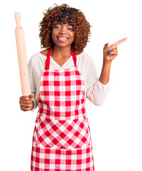 Sticker - Young african american woman wearing apron holding rolling pin smiling happy pointing with hand and finger to the side