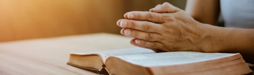 Close up of a woman hands praying on the open holy  bible on a table indoor with the windows light lay warm tone . Christian faith and trust concept  with copy space. Christian devotional background.