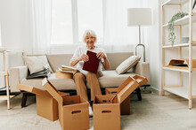 Elderly Woman Sits On A Sofa At Home With Boxes. Collecting Things With Memories And Moving And Happiness Smile.