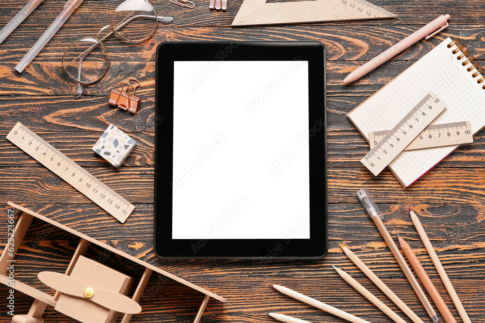 Tablet with toy plane and different stationery on brown wooden background
