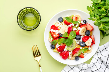 Summer fruit and berry salad with feta cheese, fresh strawberries, blueberries, banana, kiwi, orange and mint, green background, top view