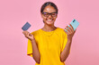 Young laughing Indian woman holding phone and credit card from bank to pay for subscriptions to mobile online cinemas or music services stands on plain pink background. Buying, shopping, banking