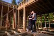 © anatoliy_gleb - Man and woman inspecting their future wooden frame house nestled near the forest. Youthful couple at construction site in early morning. Concept of contemporary ecological construction.
