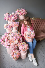 Wall Mural - a happy young girl holds a box of flowers in her hands and looks away. A beautiful woman is sitting on a sofa surrounded by flowers. A box with peonies and bush spray roses in a peach-pink range.