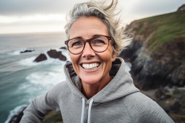 Wall Mural - Close-up portrait photography of a happy mature woman wearing a comfortable hoodie against a dramatic coastal cliff background. With generative AI technology
