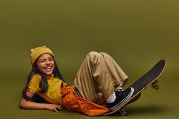 Excited and cheerful preteen girl with dyed hair wearing trendy clothes and hat while lying near skateboard and looking at camera on khaki background, girl in urban streetwear concept