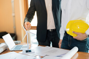 White energy wind turbine model on the desk Hands of two Asian male architects are pointing, holding helmets. And there is a blueprint about the design of the construction industry in the office.