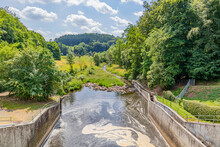 Water Flowing From Staumauer Bitburger Dam Into Prum River, Valley And Mountains With Leafy Trees In Background, Brown Spot On Water Surface, Stausee Bitburg Reservoir, Sunny Spring Day In Germany