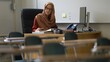 © Robert Peak - Happy woman teacher wearing headscarf in empty school classroom working on assignments for students.