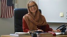 Closeup Portrait Of Happy Smiling Woman Teacher Wearing Headscarf In School Classroom Desk Gives Thumbs Up With US American Flag Behind Her.