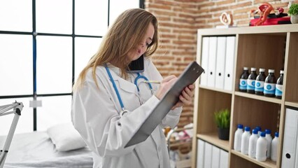 Sticker - Young blonde woman doctor writing document on clipboard talking on smartphone at the clinic