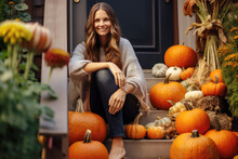 Happy Young Woman Sitting On The Stairs On A Porch Of Her Home, Beautifully Decorated With Autumn Pumpkins 