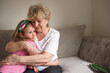© Elena - Grandmother hugging grand kid on couch at home, holding little girl in arms. senior grandma enjoying leisure time with cute granddaughter, talking to child. Family relations