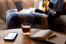Notebook, Tea And Smartphone On Table, With Woman Playing Guitar And Laptop Behind, Copy Space