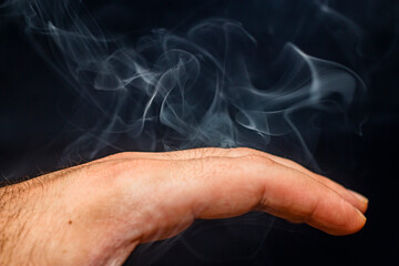 Beautiful incense smoke with beautiful bizarre shapes in the air wraps around a male palm close-up on a black background	
