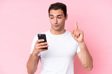 Wall Mural - Young caucasian man isolated on pink background using mobile phone and lifting finger