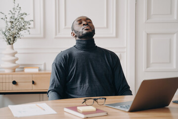 Tired Afro American employee rests, removes glasses, closes eyes, and leans back, recovering from remote work.