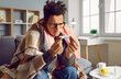 Portrait of a sick young african american man in glasses checking his temperature wrapped in a blanket on the sofa. Guy with seasonal flu or cold feel unhealthy with influenza and runny nose at home.