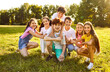 © Studio Romantic - Portrait of a happy smiling children friends sitting on the green grass together in the park outdoors and looking cheerful at the camera hugging and having fun. Kids summer camp and holidays concept.