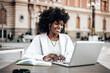 © Dusko - Beautiful Black female student sitting at the coffee bar table and using her laptop computer for work and learning. Bring sunny day. Old city buildings in the background.