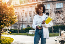 Beautiful And Happy Black Female Student Enjoying Outdoors In College Campus. She Is Happy And Smiling While Holding Laptop Computer And Walking Through College Park. Old Building In Background.
