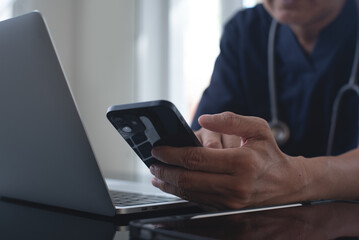 Wall Mural - Doctor using mobile smart phone, working on laptop computer in medical workspace office with stethoscope on desk, close up, teleconference, telemedicine, medical research and technology concept