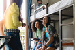 © Lomb - Multicultural Hostel Room Gathering with Smartphone - Two women, Caucasian and Brazilian, laughing with a smartphone, surrounded by friends in a hostel, boy on upper bunk laughs