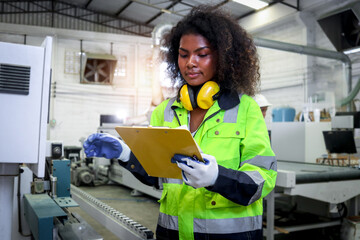 Wall Mural - African industrial woman worker with curly hair wears safety vest, writes on file folder document, female engineer check and maintenance machinery at CNC woodworking manufacturing furniture factory.