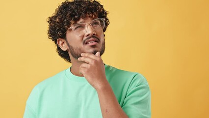 Wall Mural - Portrait of a handsome curly-haired young man in a turquoise t-shirt and glasses standing on a yellow background and thoughtfully scratching his chin.The emotion of doubt, reflection and confusion.